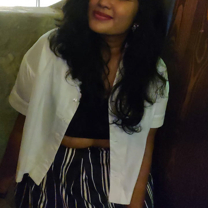 A social media shot featuring the Tanvi button up collared crop top in white. Styled over a black bandeau with a striped pant.